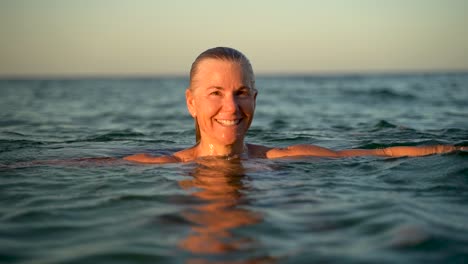Slow-motion-of-pretty-mature-woman-rising-up-through-the-water-and-smiles-at-the-camera-at-sunset