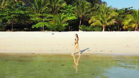 Young-woman-in-bikini-walking-on-white-sandy-beach-washed-by-calm-clear-water-of-sea-reflecting-palm-trees-on-shore-of-tropical-island-in-Ko-Pha-Ngan