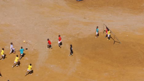 Young-children-kick-football-around-playing-soccer-on-red-dirt-pitch,-missing-shot