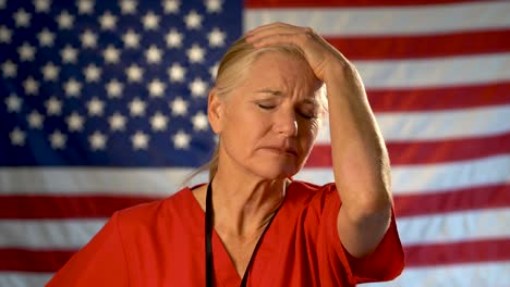 Retrato-Medio-Apretado-De-Una-Enfermera-Que-Se-Ve-Muy-Preocupada-Y-Triste-Frotándose-La-Frente-Con-La-Bandera-Estadounidense-Detrás-De-Ella