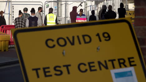 People-in-protective-face-masks-queue-up-outside-an-appointment-only-Covid-19-testing-centre-behind-a-yellow-Covid-19-Testing-Centre-street-sign