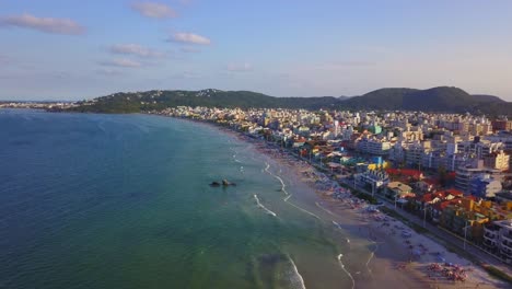 Aerial-wide-shot-of-Bombas-beach-crowded-with-tourists-at-golden-hour,-Brazil
