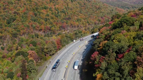 Tractor-trailers-travel-on-interstate-highway-that-curves-through-mountain-terrain-during-fall,-colorful-autumn-foliage-leaves-accent-scene