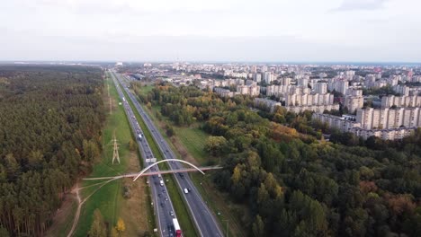 Drohne-Fliegt-Langsam-über-Die-Autobahn-A1-In-Richtung-Des-Wohnviertels-Der-Stadt-Kaunas,-Mit-Starkem-Verkehr-Und-Einer-Fußgängerbrücke-In-Der-Ferne