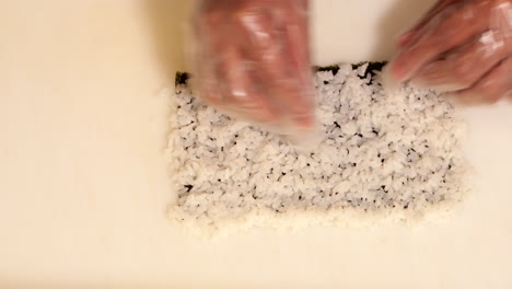 Hands-With-Disposable-Plastic-Gloves-Preparing-Sushi-Rice-On-Nori-For-Rolling---overhead-shot,-slow-motion