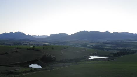 Aerial-drone-over-green-vineyards-and-ponds-with-blue-mountains-and-farmhouse-shed-in-background,-before-sunrise,-Stellenbosch