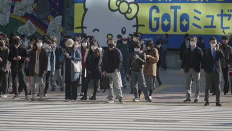 Personas-Con-Máscaras-Esperando-Para-Cruzar-La-Calle-Con-Vehículos-En-Primer-Plano-Conduciendo-En-El-Cruce-De-Shibuya-Durante-La-Pandemia-En-Tokio,-Japón