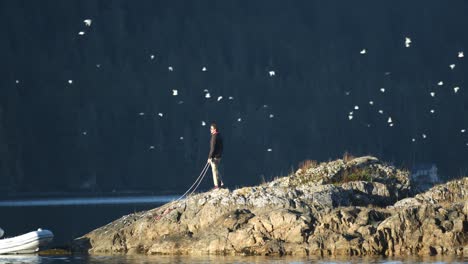 Lonely-Man-Standing-on-a-Rock-by-the-Sea-Holding-Boat-Ropes-While-Flock-of-Birds-Flying-by-the-Coast-on-Sunny-Day,-Slow-Motion