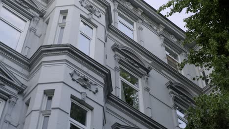 Grey-facade-of-a-beautiful-European-residential-building-in-victorian-style-from-low-angle,-with-green-tree-foliage-growing-at-the-wall