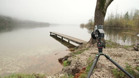 Camera-set-up-on-a-tripod-really-low-to-the-ground-on-a-foggy-day-nears-the-edge-of-a-lake-with-a-wooden-pier-in-the-background
