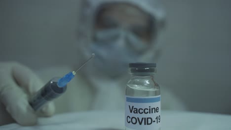 human-wearing-protective-PPE-kit-carefully-spilling-out-medicine-from-a-syringe-filled-with-covid-19-vaccine