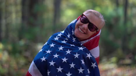 Closeup-of-blonde-woman-looking-at-camera-and-raising-an-American-flag-behind-her-with-smiling-expression-and-wrapping-the-flag-around-her-head-and-body