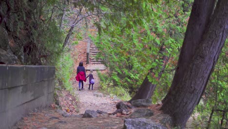A-mother-and-daughter-walk-down-a-forest-trail-in-the-late-fall-with-leaves-all-over-the-place-beside-a-green-tree-and-orange-leaves-with-a-lake