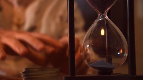 hourglass-marking-the-time-with-its-blue-sand,-with-a-man-using-tarot-cards-in-the-background