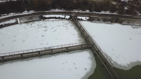 Frozen-lake-in-winter,-aerial-drone-shot-from-above,-concrete-walkways-in-lake,-dark,-cloudy-and-moody-scene,-patches-of-ice-on-water