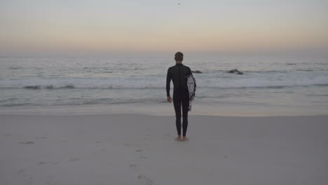 Surfer-standing-on-beach-at-sunrise-holding-surfboard-in-Cape-Town-South-Africa
