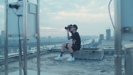 Slow-motion-open-shot-of-young-asian-photographer-capturing-urban-city-downtown-while-sitting-on-the-rooftop-in-the-early-morning