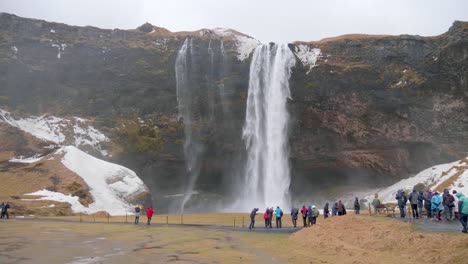 Static,-wide-shot,-of-lots-of-people-standing-in-front-and-enjoying-the-view-of-seljalandfoss-waterfall,-on-a-cloudy,-autumn-day,-in-Iceland