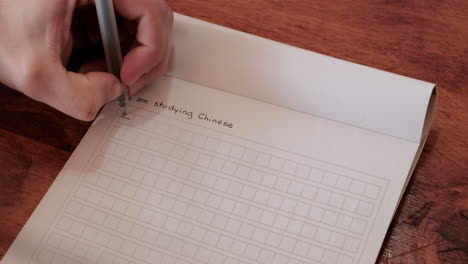 Männerhände-Schreiben:-Ich-Lerne-Chinesisch-In-Mandarin-Chinesisch-Schriftzeichen-In-Einem-Notizbuch-über-Einem-Holztisch