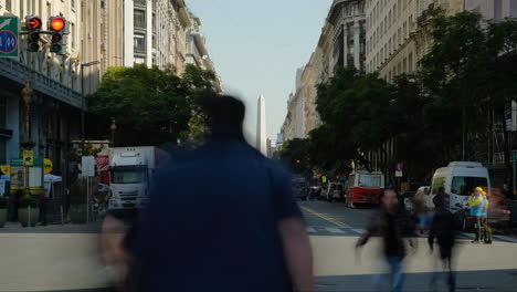 People-crossing,-public-transport-and-Obelisk-of-Buenos-Aires-at-daytime-wide-shot-timelapse-zoom-in