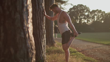 Man-In-Park-Stretching-Out-His-Legs-Before-He-Goes-For-a-Run-Whilst-Sun-Sets-Behind-Him
