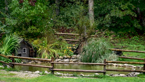 Decorative-pond-with-watermill,-pond-surrounded-with-flowers-and-gras,-wooden-fence-built-around,-steady-shot