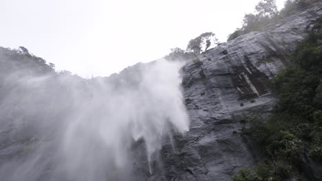 Zeitlupenaufnahme-Eines-Wasserfalls,-Der-Eine-Klippe-Hinunterstürzt-Und-Vom-Wind-Weggeblasen-Wird