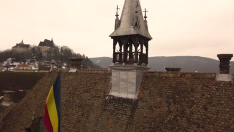 A-rotating-drone-shot,-capturing-the-rooftop-of-a-vintage-architechture-in-the-city-of-Sighisoara-on-an-afternoon-with-a-community-flag-hoisted-beside-and-a-cityscape-in-the-background