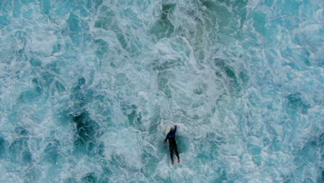 AERIAL:-Birdseye-view-of-surfer-battling-the-waves,-current-and-white-wash-in-a-turbulent-ocean