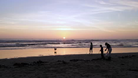 A-mother-helps-to-clean-up-trash-on-the-beach-during-sunset-at-the-beach-as-a-woman-plays-catch-with-her-dog