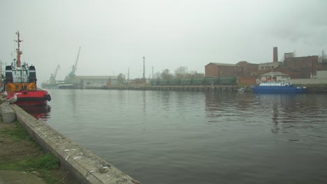 Iceboat-and-blue-fisherman's-ship-docked-at-Port-of-Liepaja-in-foggy-day,-dry-cargo-wagons-and-port-cranes-in-background,-wide-shot
