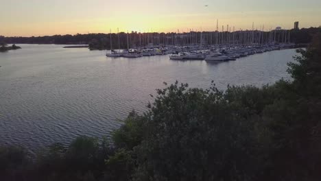 Aerial-Sunset-Descending-Shot-Of-Sailboat-Marina-Yacht-Club-Dock-In-Lake-Bay-Slides-Left-Among-Green-Trees-With-City-Buildings-Skyline-In-Background-In-Toronto-Ontario-Canada