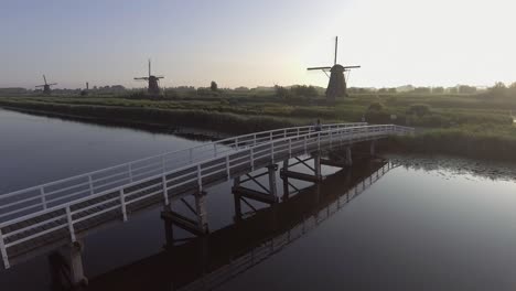 A-drone-shot-panning-left,-around-a-girl-walking-and-turning-around-on-a-bridge,-looking-at-Dutch-Windmills-in-the-Netherlands-during-sunrise