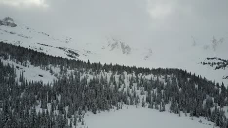 Drohne-Fliegt-Im-Winter-über-Den-Zugefrorenen-See-Und-Nähert-Sich-Der-Baumbedeckten-Bergkette