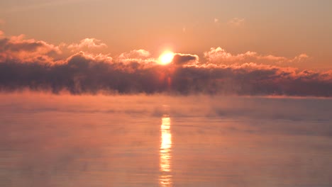 Timelapse-of-the-sun-rising-through-clouds-over-a-very-cold-lake-with-steam-rising-in-the-winter-time-as-red-yellow-and-red-sun-rays-brighten-the-sky