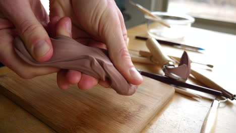 Slide-across-the-hands-of-an-artist-sculpting-with-brown-modeling-clay-on-a-wooden-table-with-tools-strewn-about-the-art-studio
