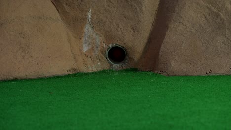 An-orange-mini-golf-ball-exits-a-pipe-in-a-wall-and-rolls-over-for-a-hole-in-one-shot