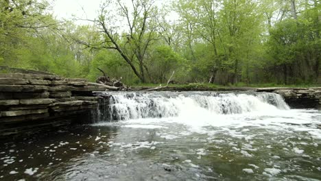 Low-to-the-water-surface-moving-in-and-going-above-a-small-forest-river-waterfall-in-a-rural-park