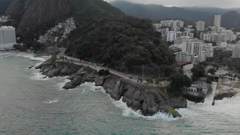 Aerial-approach-on-Leblon-viewpoint-and-parking-with-waves-crashing-in-on-the-rocky-coast-on-a-gray-overcast-day-in-Rio-de-Janeiro