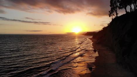 Aerial-drone-shot-of-an-orange-sunset-sky-above-the-ocean-waves-off-the-sandy-beach-cliffs-of-Santa-Barbara,-California