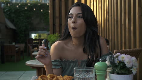 Adorable-woman-eating-curly-fries-in-a-cafe-in-London,-hispanic-latina-millennial-at-the-table-in-a-casual-outfit,-eating-and-having-fun,-smiling-and-enjoying-the-fast-food