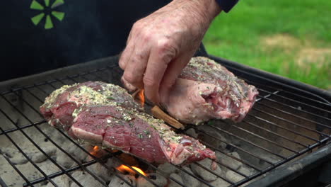 Two-pieces-of-lamb-meat-are-being-grilled-on-a-coal-fired-BBQ,-person-is-adding-wood-chips-for-flavor