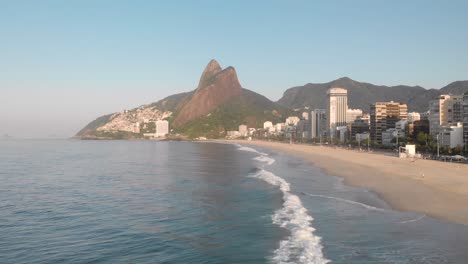 Aerial-backing-up-above-the-ocean-waves-with-a-view-of-coastal-city-beach-of-Rio-de-Janeiro-during-early-morning-golden-hour