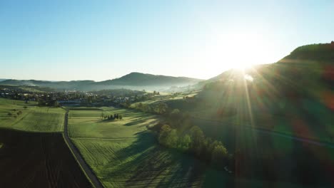 aerial-drone-view-of-morning-hour-over-peaceful-countryside-with-green-morning-dew-and-agricultural-fields,-sun-shining-directly-into-camera,-switzerland