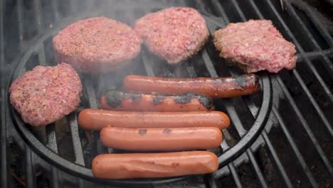 outdoor-grill-closeup-as-seasoned-beef-hamburger-patties-and-hot-dogs-are-turned-over-flames-with-smoke