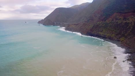 Flying-over-turquoise-ocean-waves-with-yellow-sand-cloud-in-the-water-towards-a-Coastal-Highway-bridge-in-Big-Sur-California
