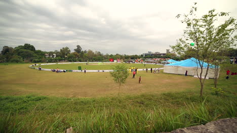 A-sports-ground-long-panning-view,-grass,-trees-and-camps-around-the-ground,-People-are-outside-and-inside-of-the-ground