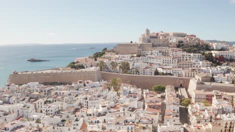 Aerial-view-of-Ibiza-city,-the-Old-Town-and-the-city-walls-of-Eivissa,-in-the-island-of-Ibiza,-on-a-sunny-and-clear-day,-just-after-sunrise,-with-birds-flying-alongside-the-city-wall
