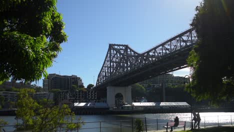 Frühmorgendliche-Spaziergänger-Schlendern-Am-Ufer-Des-Sonnendurchfluteten-Brisbane-River-Am-Kangaroo-Point-Entlang,-über-Dem-Sich-Die-Berühmte-Story-Bridge-Hoch-Erhebt