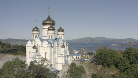 Slow-descent-aerial-shot-of-seaside-orthodox-church-with-blue-roof-and-golden-domes-with-port-and-bay-in-the-background-on-a-bright,-sunny-day